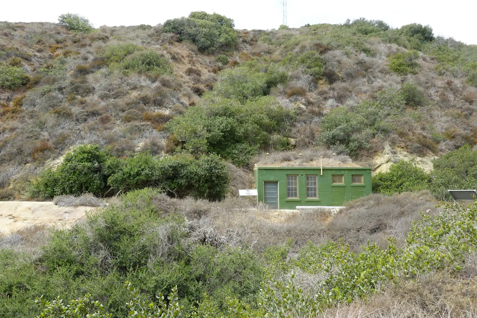 A small building at the foot of a hillside covered in dense, dry brush